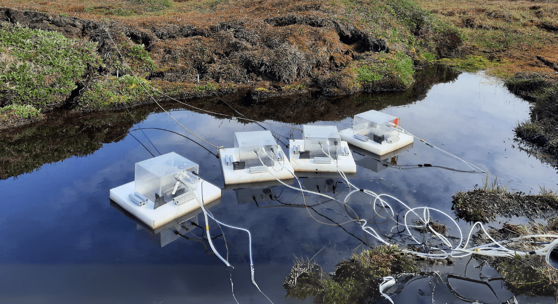 Floating scientific equipment with wires is set up on a reflective body of water, surrounded by grassy and mossy terrain, indicating a research setting.