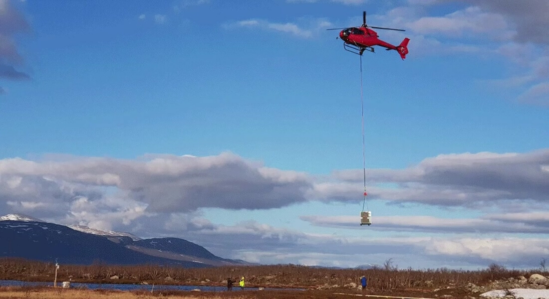 A red helicopter hovers in a blue sky, lifting a large crate with a cable over a mountainous, partly snowy landscape.