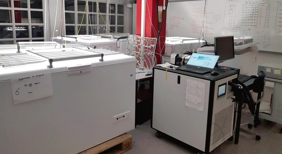 A laboratory with white freezers containing scientific equipment. A computer station is nearby, displaying data. 